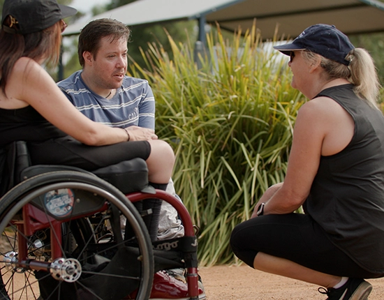 2 Ability West clients in wheelchairs while Cassie crouches to speak with them