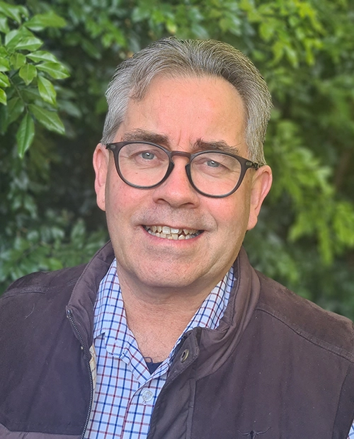 Mark Gardner is standing in front of a hedge, wearing a blue and white check shirt and vest